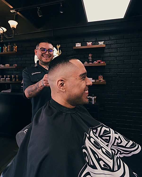 Barbero de lentes trabajando un fade, con repisas de productos en el muro de ladrillo negro