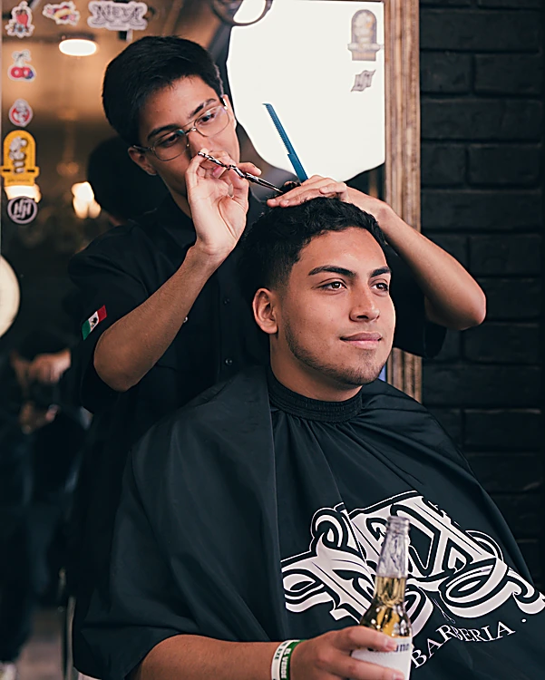 Barbero cortando con tijera y peine a un cliente sonriente con capa de MEXA, frente al espejo de marco dorado