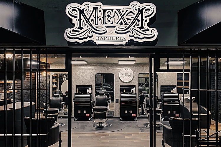 Fachada de La Mexa de noche: el letrero MEXA Barbería sobre la entrada y, detrás de la reja, cuatro sillones negros frente a sus espejos