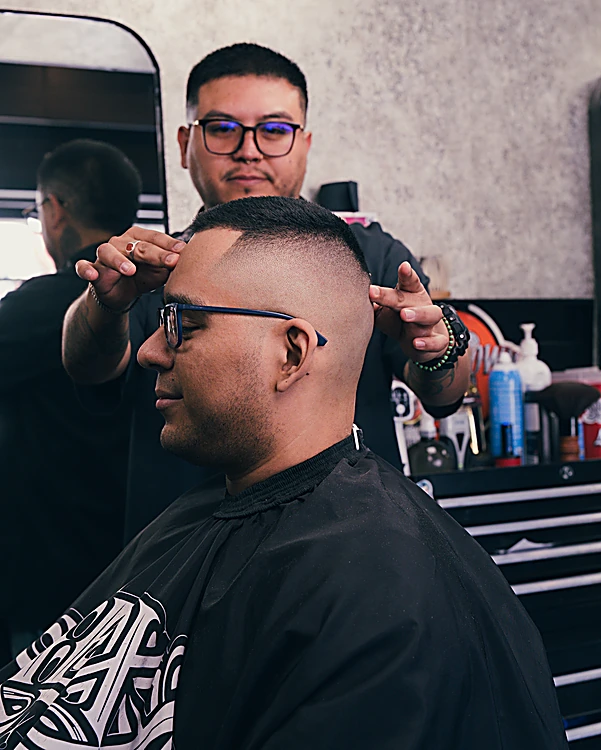 Barbero de lentes acomodando la cabeza de un cliente con fade alto a máquina, capa negra con la filigrana de MEXA