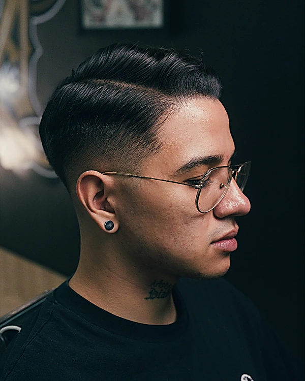 Perfil de un cliente con lentes redondos: fade bajo en los lados y el cabello de arriba peinado hacia un lado