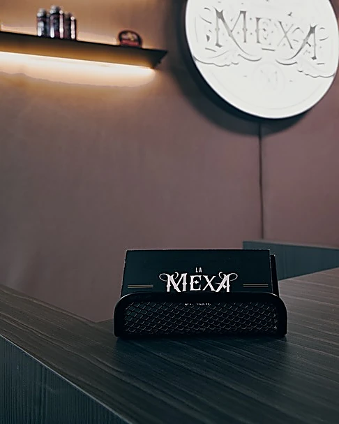 Toalla negra doblada con el logo MEXA sobre la barra de madera oscura, bajo una repisa con luz y el letrero redondo